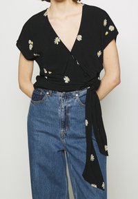 Blusa nera avvolgente con maniche corte, caratterizzata da ricami di margherite bianche e gialle. Abbinata a jeans in denim blu a vita alta.