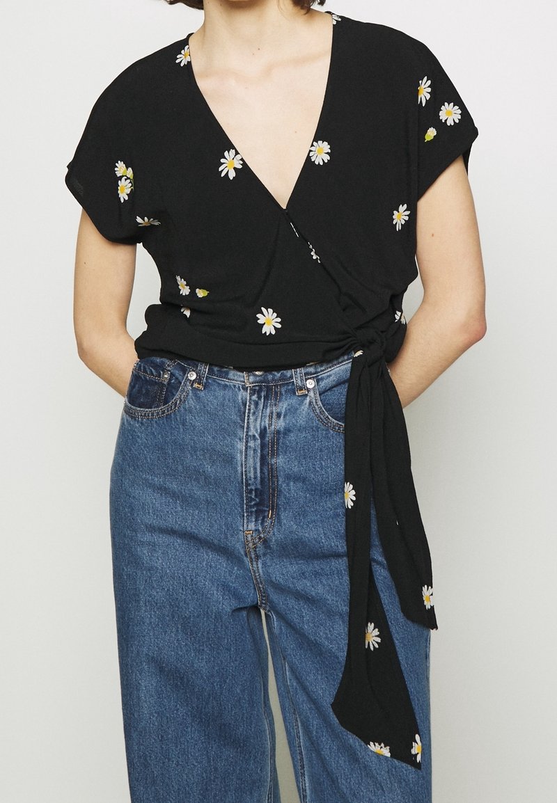 Blusa nera avvolgente con maniche corte, caratterizzata da ricami di margherite bianche e gialle. Abbinata a jeans in denim blu a vita alta.