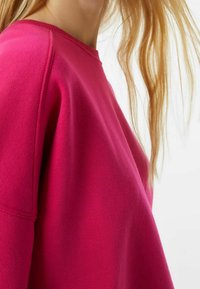 Sudadera fucsia con cuello redondo, hombros caídos, textura suave y corte relajado. Detalle de costura visible en las mangas.
