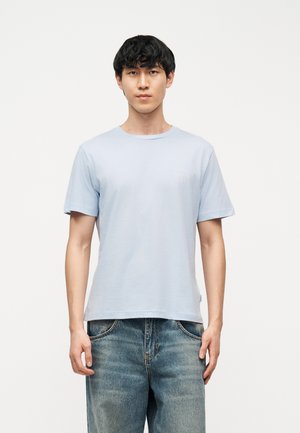 Mladý muž s čiernymi vlasmi, oblečený v svetlomodrom tričku s krátkym rukávom a vyblednutých modrých džínsoch, stojaci pred jednoliatym bielym pozadím.