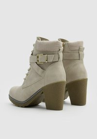 Botas de tobillo beige con un cuello texturizado y acanalado, cierre frontal con cordones, cremallera lateral y tacón grueso. Acentuadas con un detalle de hebilla dorada.