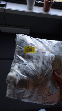 Bela in modra črtasta srajca, zložena in pakirana v prozorno plastiko, z rumeno ceno in več oznakami vidnimi na embalaži.