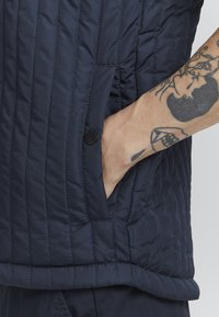 Jūrų karinės mėlynos spalvos g quilted liemenė su vertikaliomis siūlėmis, kuri turi šoninį kišenę su juodu mygtuku ir lygiu paviršiumi. Matomi tatuiruotės ant riešo.