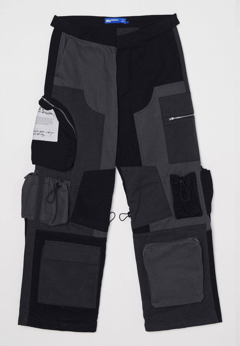 Pantalons cargo en patchwork noir et gris avec de multiples poches, fermetures éclair et détails à cordon de serrage. Fabriqués en tissu durable, avec des textures variées.