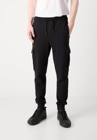 Schwarze Jogginghosen mit einem Kordelzugbund, seitlichen Taschen mit Klappen und schmal zulaufenden Beinen. Getragen mit schwarzen Sneakers auf einem neutralen Hintergrund.