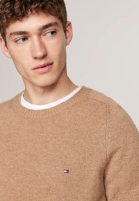 Maglione beige lavorato a maglia con collo a coste e texture sottile. Presenta un piccolo logo tricolore sul petto. Design con collo rotondo.