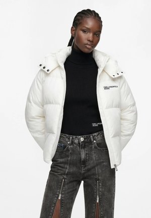 Femme portant une doudoune blanche sur un col roulé noir et un jean noir taille haute avec des fermetures éclair verticales à l'avant.