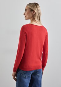 Rode gebreide trui met lange mouwen en een wijde halslijn, met ribbelmanchetten en -zoom. Het model draagt het met blauwe spijkerbroek.