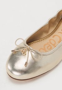 Metallisch goldene Ballerinas mit abgerundetem Zehenbereich, dekorativem Bogen mit goldfarbenen Spitzen und einem glatten Lederoberteil. Sichtbares Detail der strukturierten Innensohle.