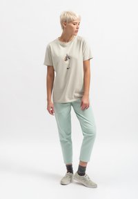 Lichtbeige katoenen t-shirt met een grafisch ontwerp, gecombineerd met mintgroene broeken. Het model draagt grijze sokken en gestreepte sneakers.