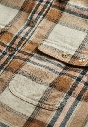 Camisa de franela a cuadros en tonos marrones, beige y negro. Presenta dos bolsillos frontales y cierres de botones. Tejido suave y texturizado con patrones de espiga.