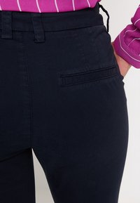 Granatowe spodnie o gładkiej teksturze, z tylną kieszenią i szlufkami na pasek, w zestawieniu z fioletowym topem w paski widocznym na wysokości talii.