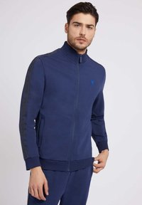 Marinefarbener Reißverschluss-Trainingsjacke mit hohem Kragen, zwei seitlichen Taschen, gerippten Bündchen und logodetail entlang der Ärmel in einer kontrastierenden Farbe.