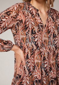 Robe à manches longues avec un décolleté en V, présentant un motif complexe de feuilles orange et blanches sur fond noir. Tissu léger et respirant.