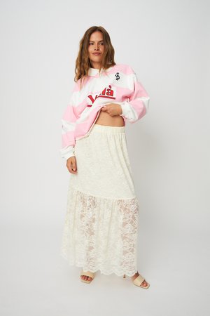 Ženska, ki nosi roza-beli pulover z navpičnimi črtami, dviguje njegov rob, pri čemer pokaže trebušček, oblečena v dolgo belo čipkasto krilo in bež sandale.