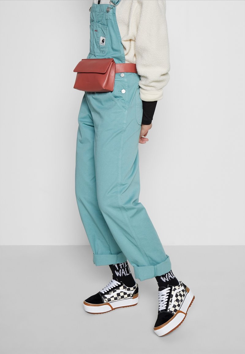 Personne portant une salopette bleu sarcelle, une veste en polaire crème, des chaussettes noires avec texte blanc, des baskets à plateforme à carreaux et un sac ceinture corail.