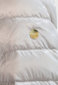 Jasnoszara kurtka puchowa z połyskującym wykończeniem, pikowaną teksturą i okrąłym złotym akcentem z logo. Widoczne linie szycia.