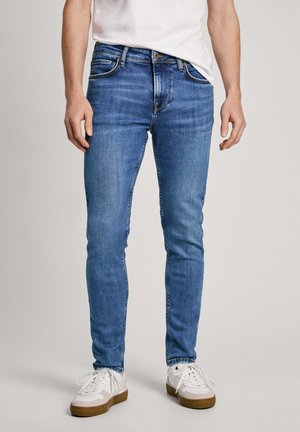 Homme portant un jean slim bleu, des baskets blanches et un t-shirt blanc, debout avec les mains détendues le long du corps sur un fond neutre.