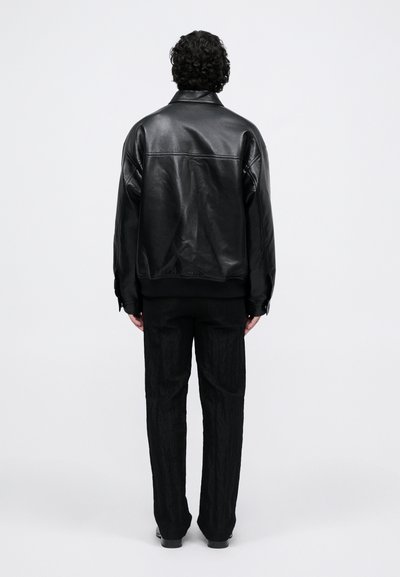 Uomo con capelli ricci che indossa una giacca di pelle nera e pantaloni neri con textura, in piedi di spalle contro uno sfondo bianco semplice.