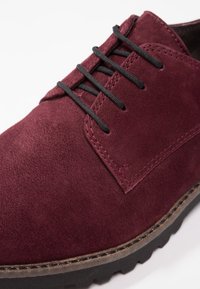 Chaussure en daim bourgogne avec lacets noirs, présentant une texture lisse et des coutures contrastées le long des bords. Semelle en caoutchouc avec des rainures.