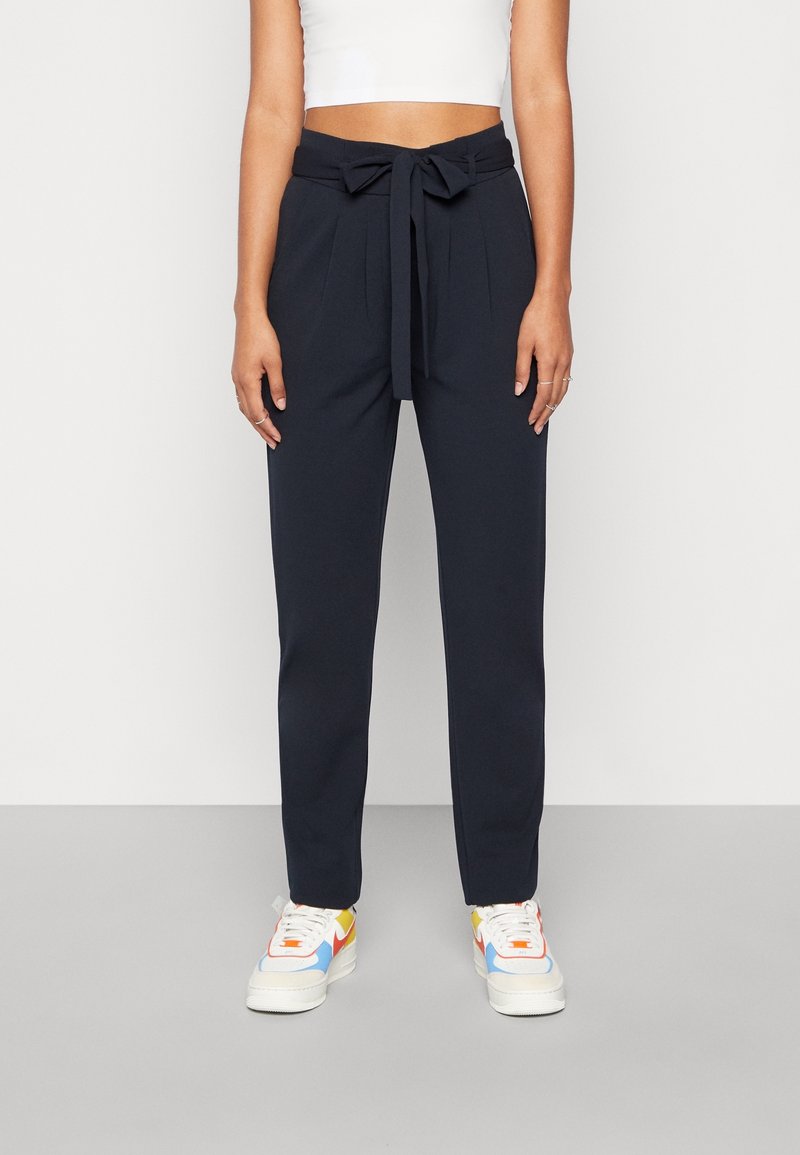 Pantalons taille haute navy avec une ceinture nouée, dotés de plis, de jambes fuselées et d'une texture lisse, associés à des baskets multicolores.