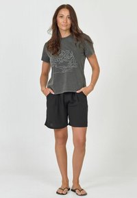 Camiseta gris de manga corta con un diseño gráfico en blanco, combinada con pantalones cortos negros. El modelo está de pie con las manos en los bolsillos, usando chanclas negras.