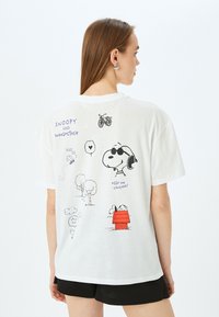Witte katoenen T-shirt met zwarte en gekleurde cartoonillustraties van Snoopy, Woodstock, bomen en tekst op de achterkant. Korte mouwen, ontspannen pasvorm.