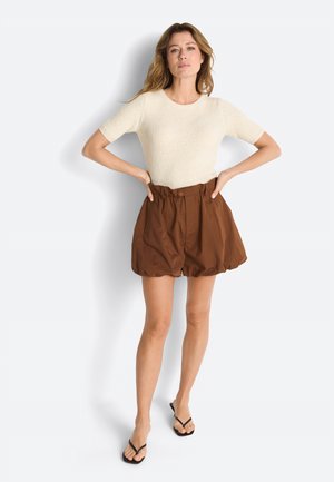 Woman with wavy blonde hair wearing cream short-sleeve knit top, high-waisted brown puff shorts, and black flip-flops, standing with hands on hips.