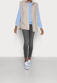 Chaleco sin mangas gris claro sobre un sudadera azul, combinado con unos jeans ajustados gris oscuro y zapatillas azul claro.