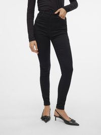 Crne skinny traperice visokog struka s glatkom teksturom i prednjim džepovima, kombinirane s crnim slingback potpeticama s oštrim vrhovima i detaljem u obliku mašne.