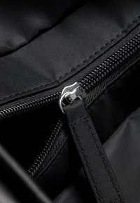 Unutrašnjost crne tkanine torbe sadrži srebrni metalni patent zatvarač i privjesak za povlačenje, s teksturiranim šavovima i glatkim rubovima.