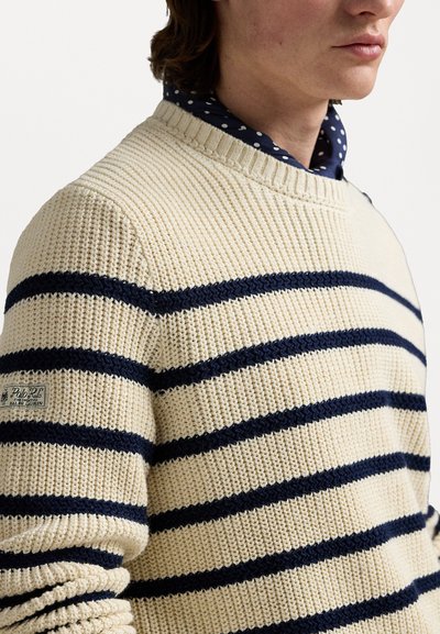 Pull en tricot crème avec des rayures navy, col rond simple, tissu texturé et une patch logo sur le bas à gauche.