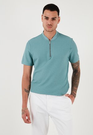 Polo majica kratkih rukava od teksturirane teal tkanine s uspravnim ovratnikom i zatvaranjem na četvrtinom patentnom zatvaraču, uparena s bijelim hlačama.
