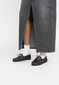 Scarpe da barca in pelle nera con suole bianche, lacci rosso-marroni e occhielli metallici, abbinate a calze bianche a scallop e una gonna di jeans grigia.