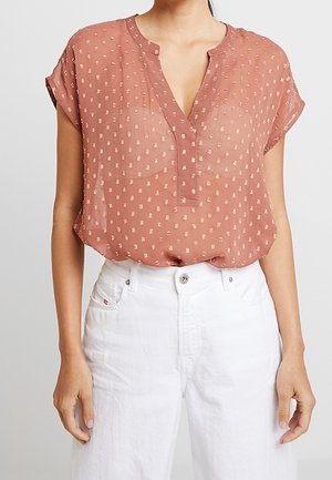 Frau trägt eine kurzärmelige, staubrosafarbene Bluse mit kleinen goldenen Mustern und weiße hoch taillierte Hose.