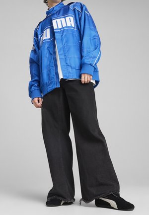 Personne portant une veste Puma bleue, un pantalon large noir avec des ourlets effilochés et des baskets noires avec des bandes blanches Puma, debout devant un fond gris.