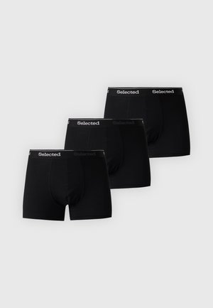 Homme musclé debout, pieds nus, portant un boxer noir avec la ceinture portant le logo "Selected", regardant vers sa droite sur un fond uni.