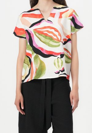 Femme portant un haut blanc à manches courtes avec des motifs abstraits rouges, verts, noirs et roses et un pantalon noir avec un lien à l'avant.