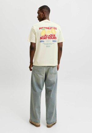 Junger Mann trägt ein cremefarbenes T-Shirt mit Zitronengrafik und rotem Text, kombiniert mit locker sitzenden hellblauen Jeans und braunen Schuhen, steht vor weißem Hintergrund.