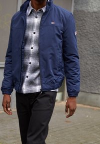 Granatowa lekka kurtka z elastycznymi mankietami, noszona na szaro-czarnej koszuli w kratę zapinanej na guziki, w połączeniu z ciemnymi spodniami.