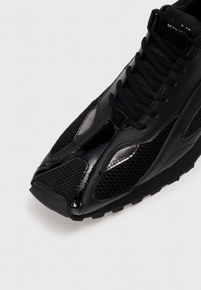 Zapatilla deportiva negra con un cuerpo de malla, acentos de cuero brillante, suela texturizada y diseño de cordones con una forma aerodinámica.