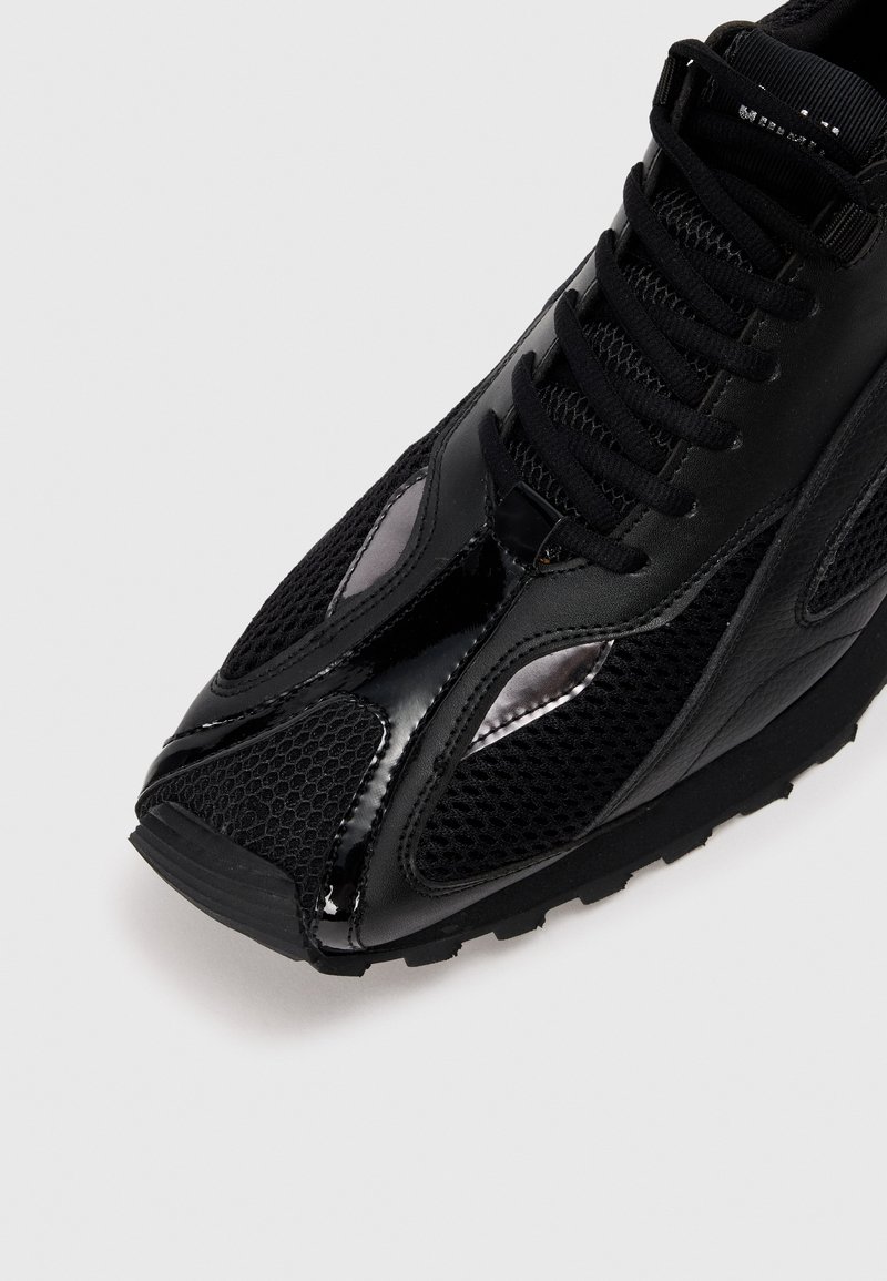 Sort sportsko med en mesh-overdel, blanke læderdetaljer, struktureret sål og snørebåndsdesign med en strømlinet form.