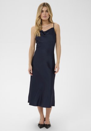 Femme aux longs cheveux blonds portant une robe midi sans manches bleu marine et des talons noirs pointus, debout sur un fond blanc uni.