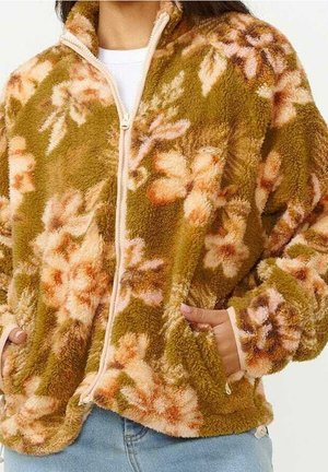 Woman wearing a fuzzy, zip-up jacket with a green and peach floral pattern, hands in pockets, paired with light blue jeans.