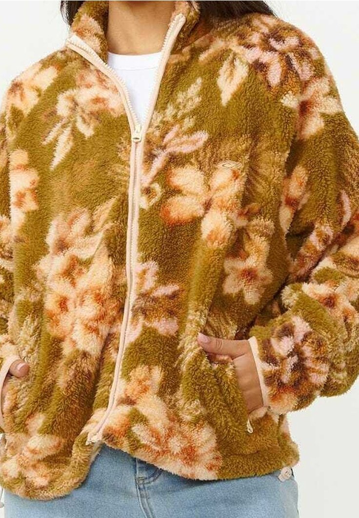 Femme portant une veste duveteuse zippée à motif floral vert et pêche, les mains dans les poches, associée à un jean bleu clair.