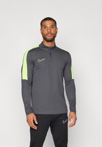 Grijze sporttop met lange mouwen en een halve ritskraag, voorzien van felgroene accenten op de schouders en een klein Nike-logo op de borst.