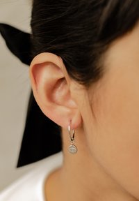 Orecchino a cerchio in argento con un piccolo ciondolo caratterizzato da un design testurizzato. Indossato su un orecchio umano, con una pelle liscia e capelli scuri.