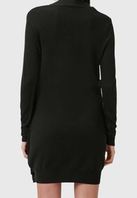 Femme portant une robe-pull en tricot noir à manches longues vue de dos sur un fond uni.