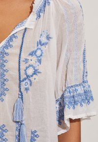 Vitt blus med blå blommönster och geometriska mönster, med en tofsuppbinding, rundad halsringning och volangärmar. Lättviktigt tyg.