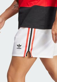 Bílé šortky s červenými a černými bočními proužky, s logem Adidas. Vyrobené z lehkého materiálu s detailem zipu na lemu.